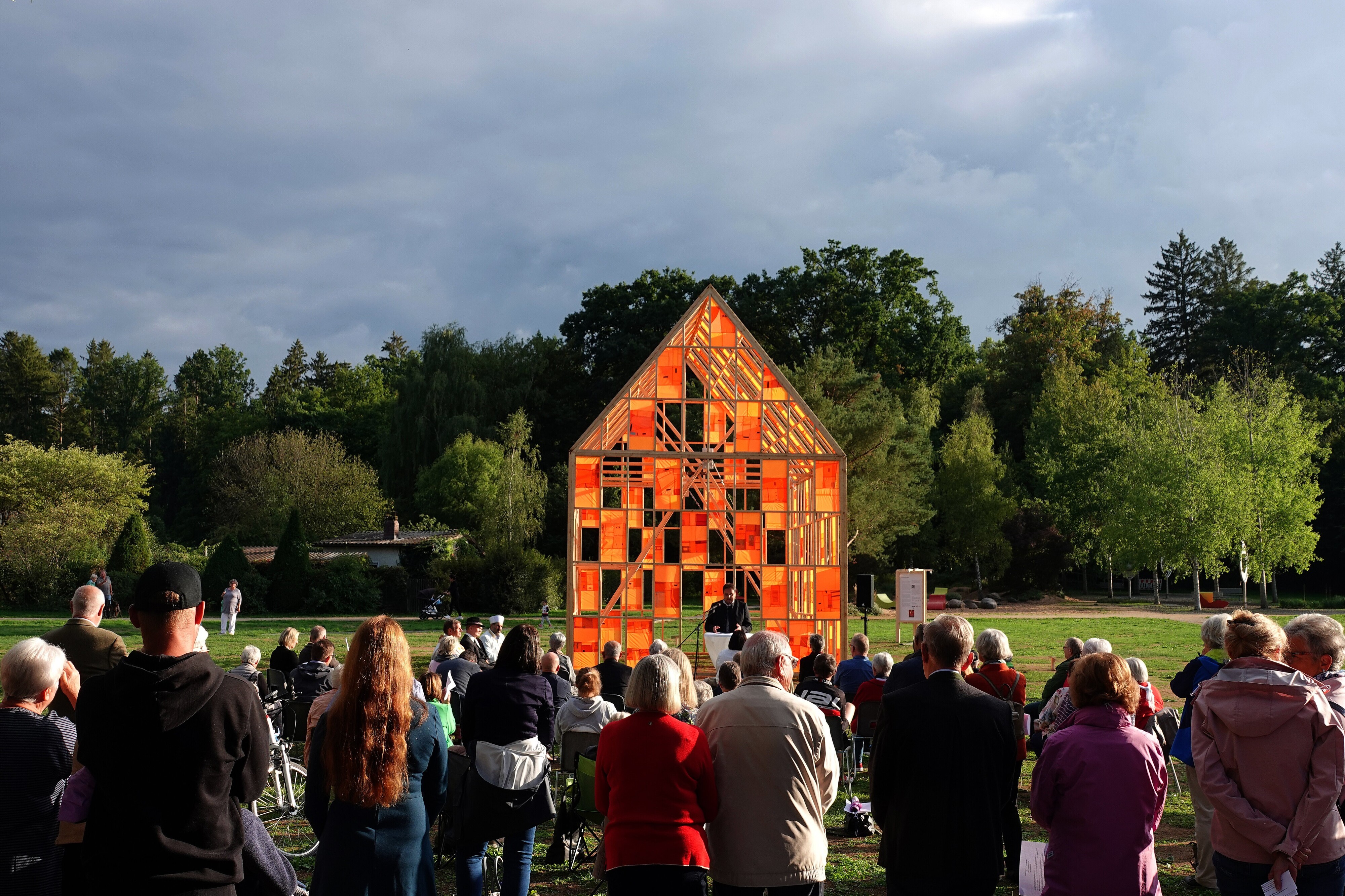 Großes Publikum steht auf der Wiese vor dem orange leuchtenden Haus