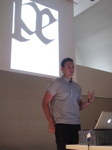 A photo of a man giving a lecture while behind him is a typographic letter on a white projected screen.