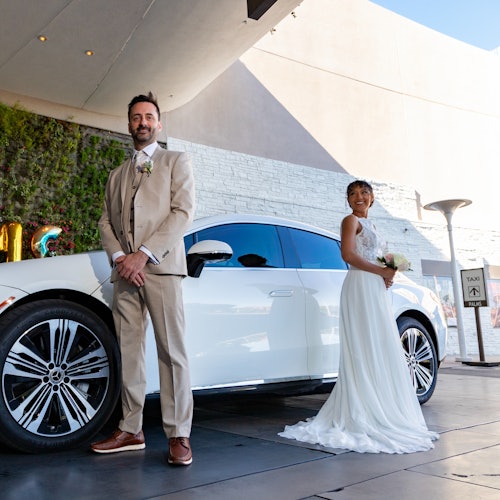 一对新娘和新郎在一辆白色汽车前摆姿势。新郎穿着米色西装,新娘穿着白色礼服,手持一束花。