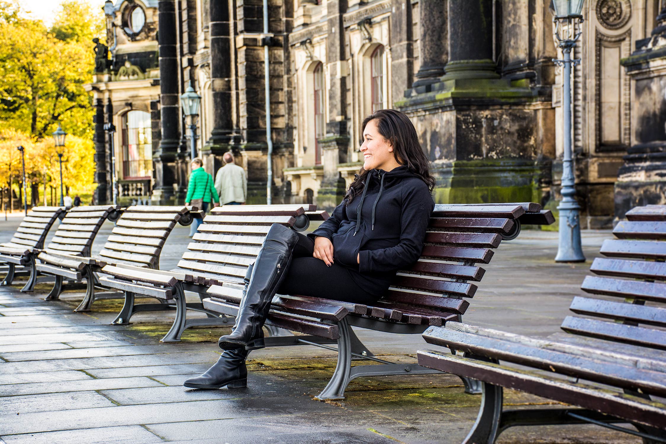A woman sits on an outdoor bench near large stone buildings while several empty benches line the wet pavement behind her.