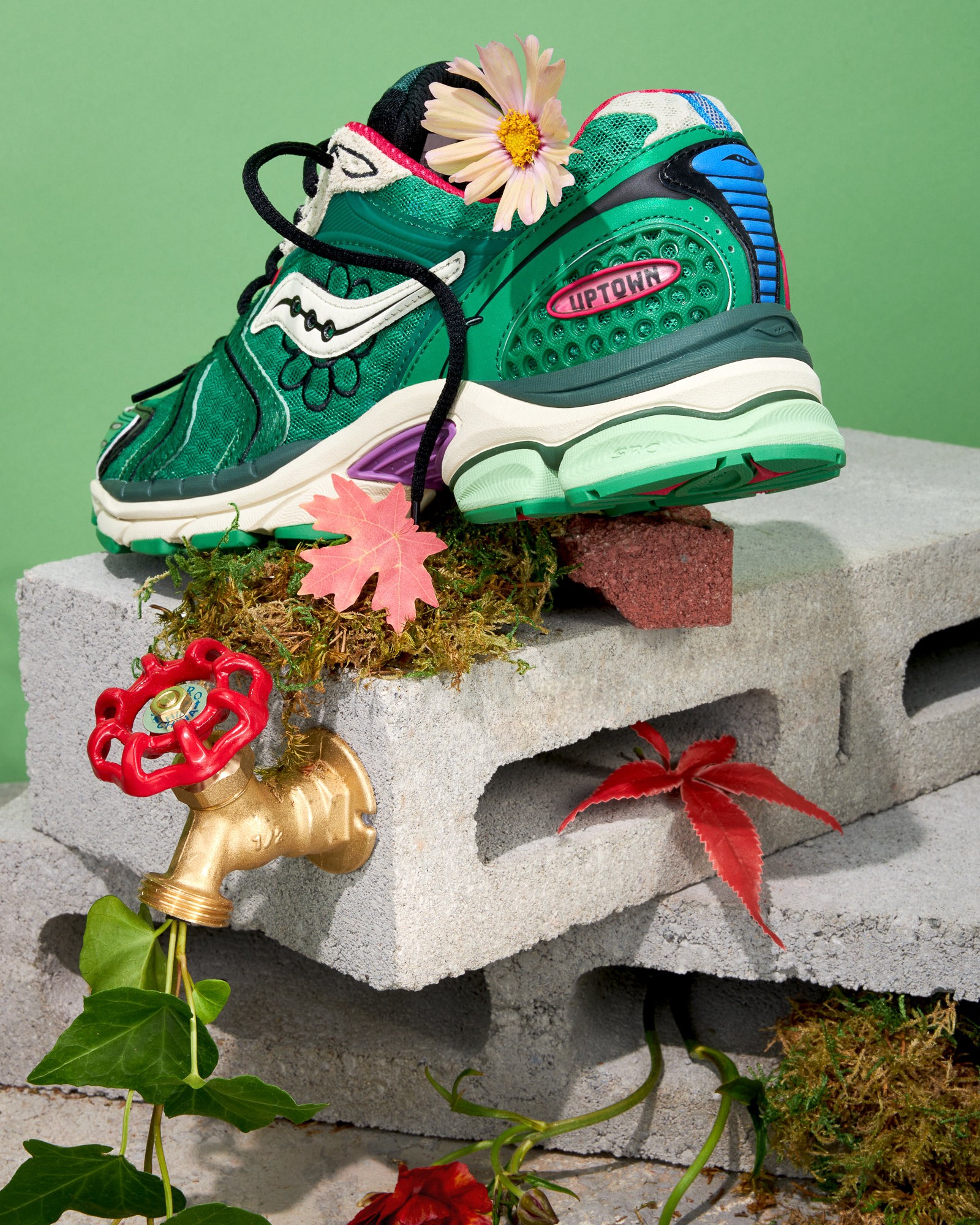 Green Jae Tips x Saucony sneaker angled on stacked blocks with flowers and garden elements in the frame.