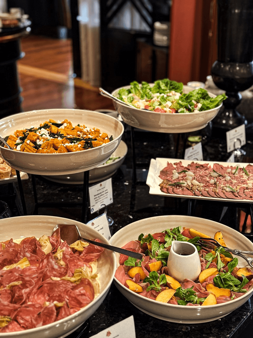 A sumptuous buffet spread features a variety of dishes including a bowl of sweet potato salad with feta and seeds, a mixed green salad with croutons, thinly sliced cured meats presented elegantly, and a vibrant antipasto platter adorned with arugula and sliced peaches.