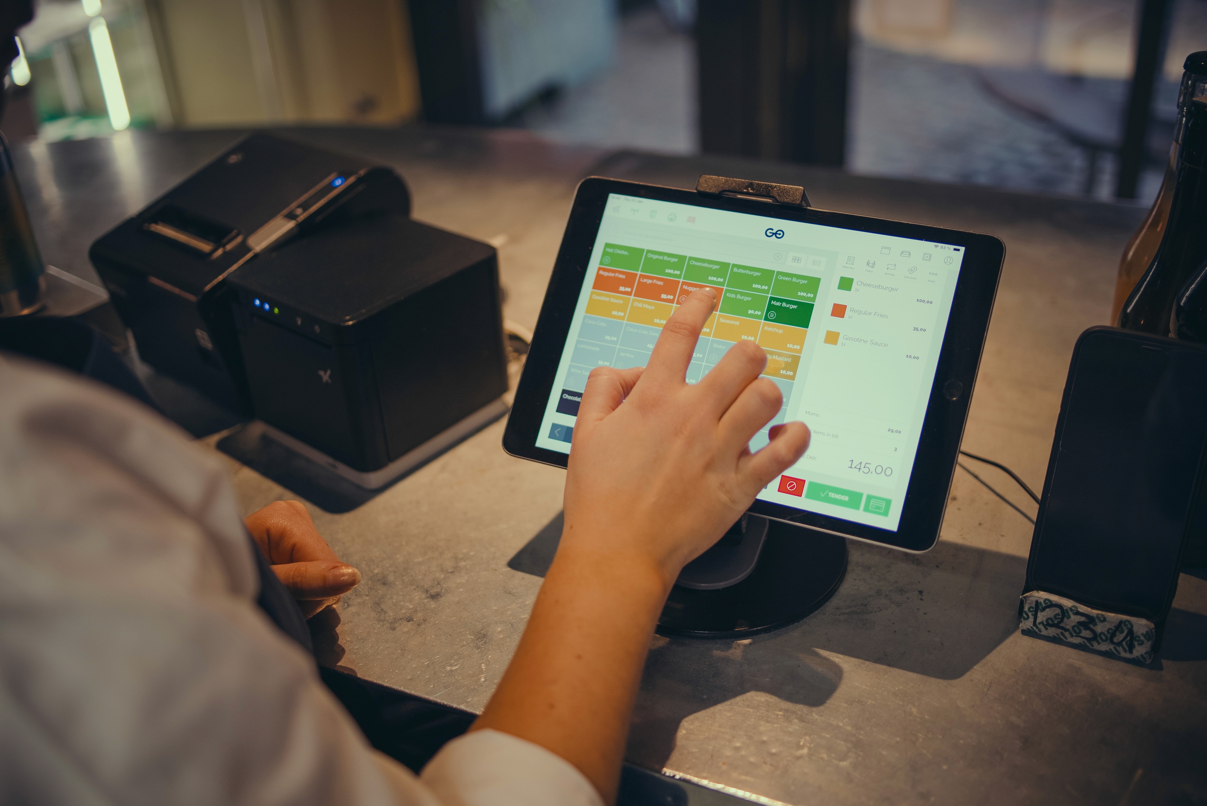 A young person with curly hair smiles at the camera while sitting in front of a laptop against a dark background.