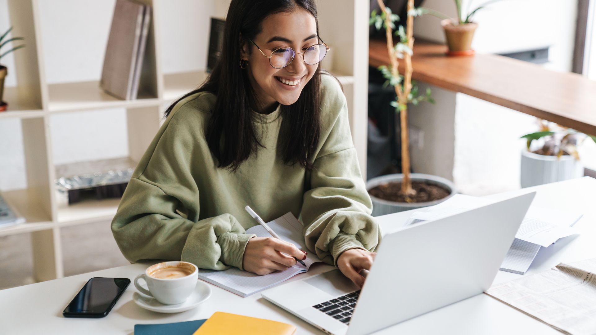 A teacher in glasses smiles while making notes beside her laptop