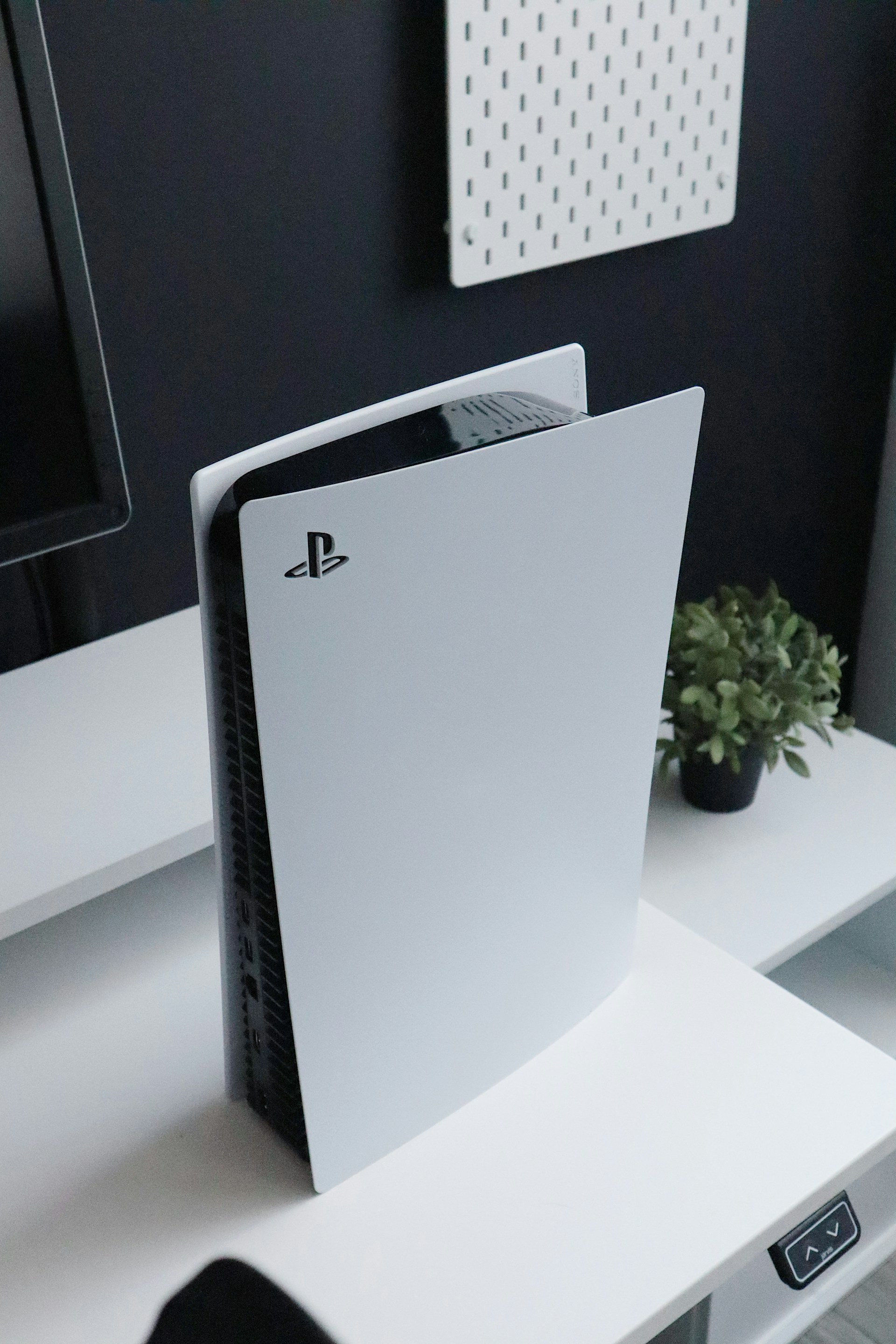 White PlayStation 5 console standing upright on a white desk next to a small potted plant.