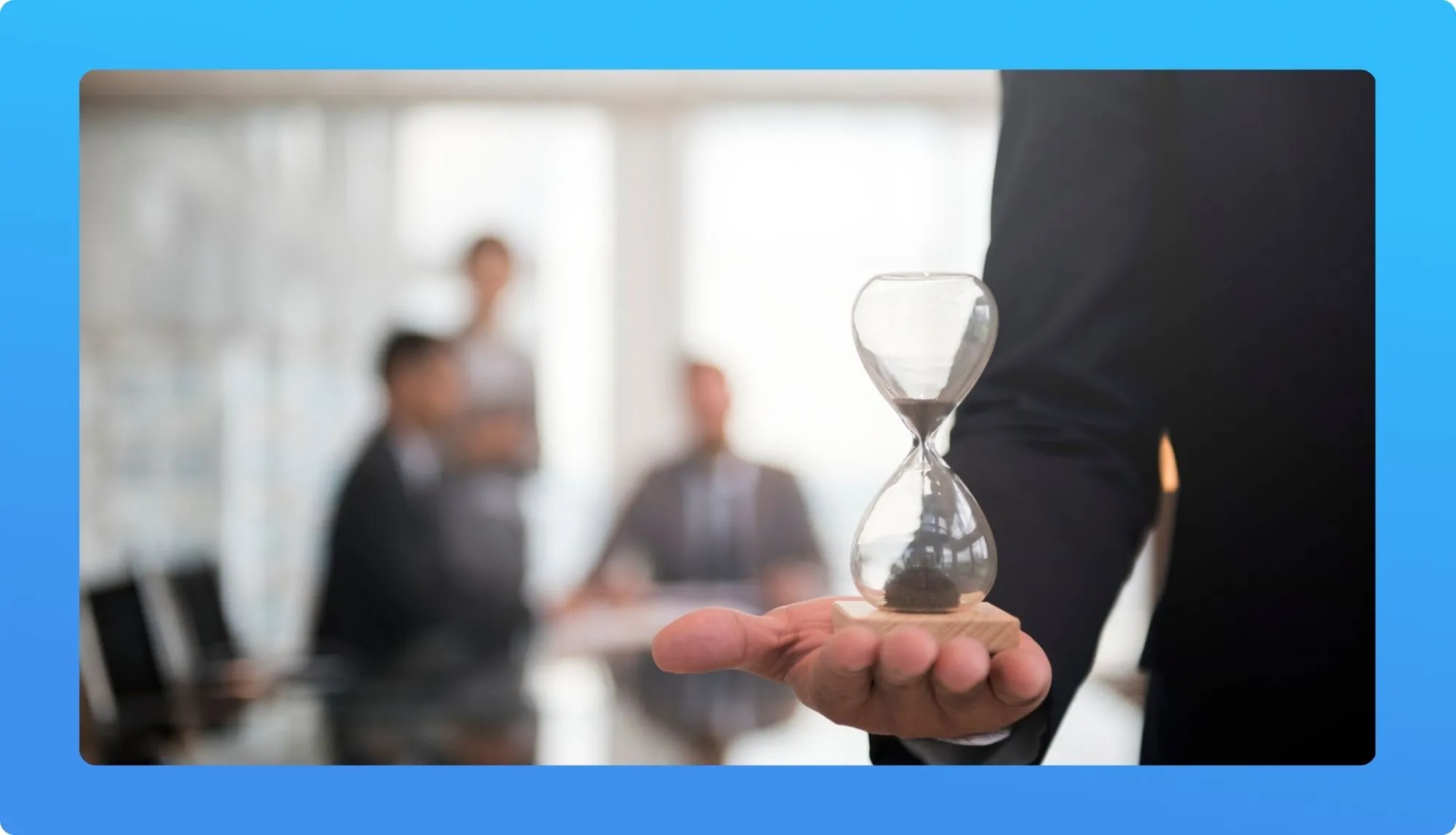A businessman holds an hourglass in his palm, symbolizing time management, with blurred colleagues engaged in discussion in the background.