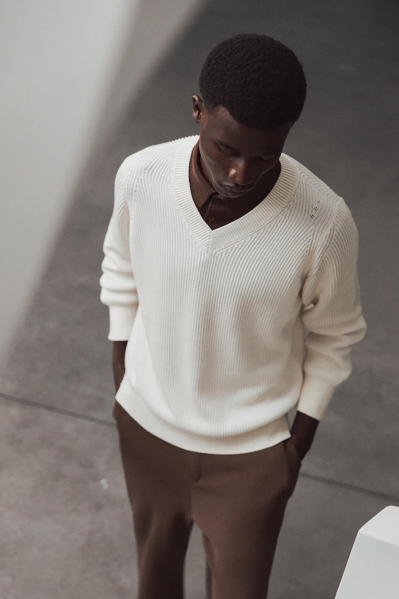 High-angle view of Aida Barni SS26 collection men's cream-colored ribbed V-neck sweater layered over a brown polo and paired with brown trousers.