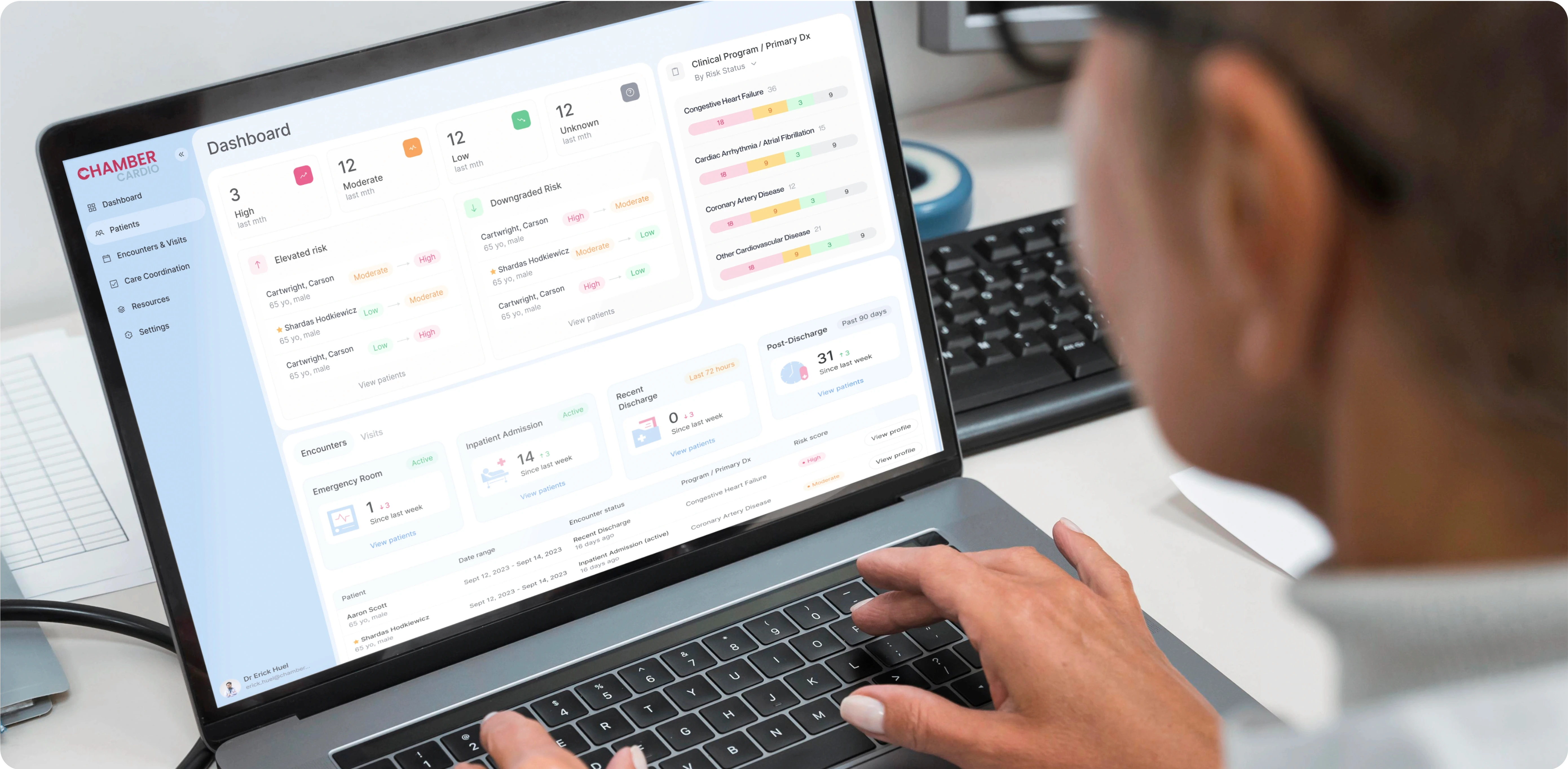 A close-up image of a person using a laptop displaying a health management dashboard. The interface on the screen belongs to ChamberCardio and is organized into various sections, including:  Risk Levels: "High," "Moderate," "Low," and "Unknown" categories for patients based on their clinical data. Encounters and Visits: Tracking recent visits to the emergency room, inpatient admissions, and discharges. Clinical Program/Primary Diagnosis: Monitoring conditions such as "Congestive Heart Failure," "Coronary Artery Disease," and other cardiovascular diseases. The dashboard provides a clear, organized overview of patient data, enabling healthcare professionals to monitor risks and patient encounters efficiently. The layout is user-friendly with soft colors, charts, and labeled sections, emphasizing the use of technology in healthcare management.