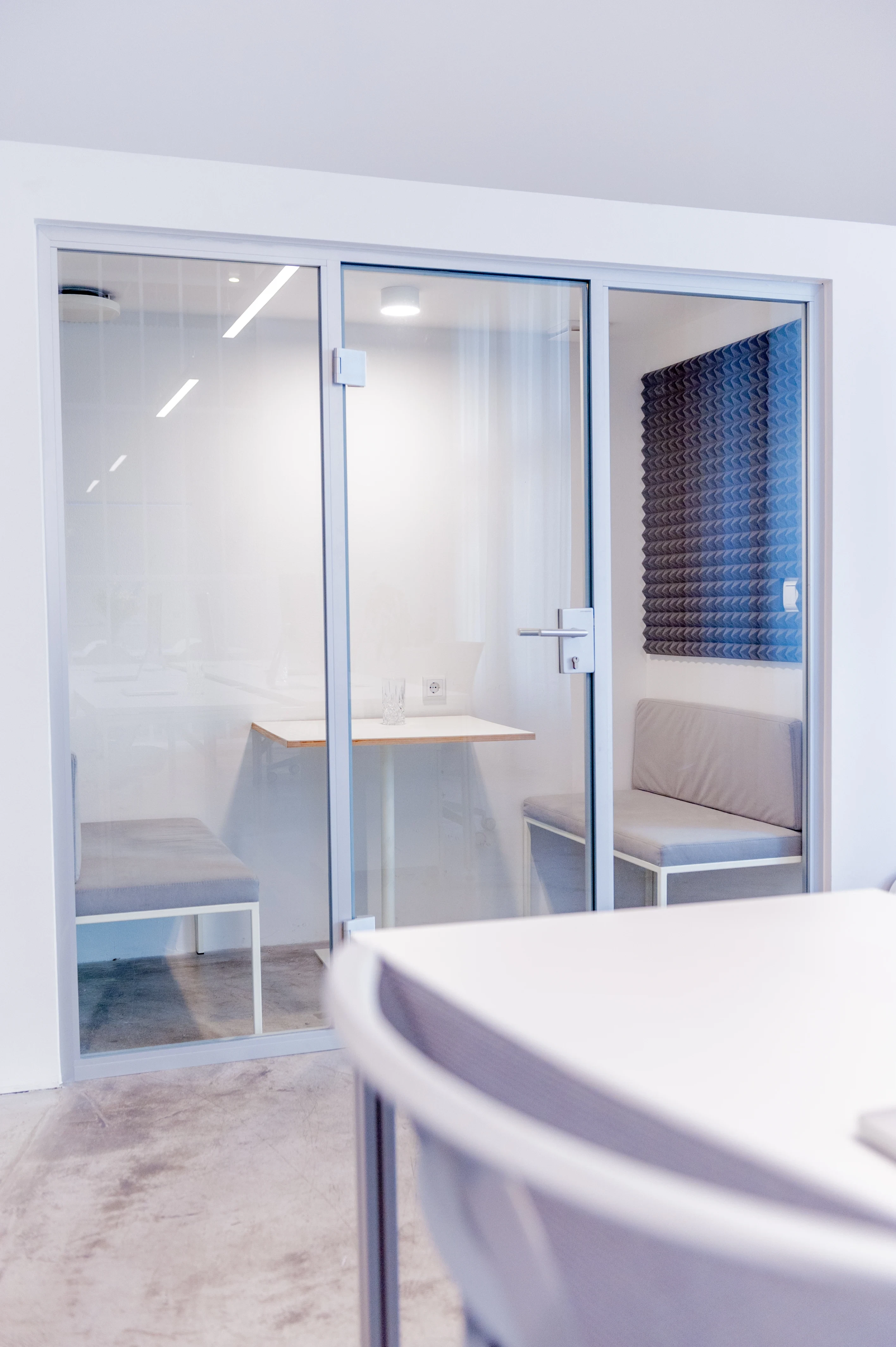 Glass-walled focus room at BATTLE ROYAL with grey upholstered benches, a small white table and a dark blue sculpted acoustic panel on the wall.