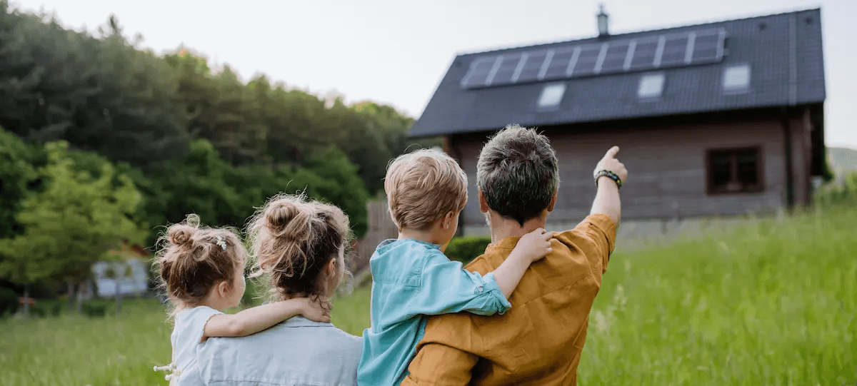 Familie (Vater, Mutter und zwei Kinder) steht vor Eigenheim und Vater zeigt auf das neue Eigenheim