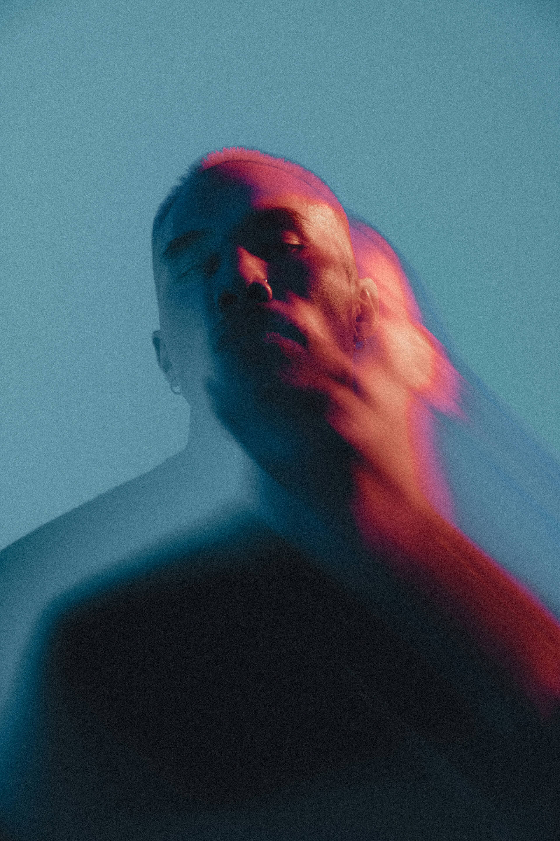 Artistic portrait of a person with a blurred motion effect, lit dramatically in blue and pink neon tones against a soft teal background. The image conveys a sense of movement, introspection, or surreal emotion through its abstract lighting and dynamic blur.