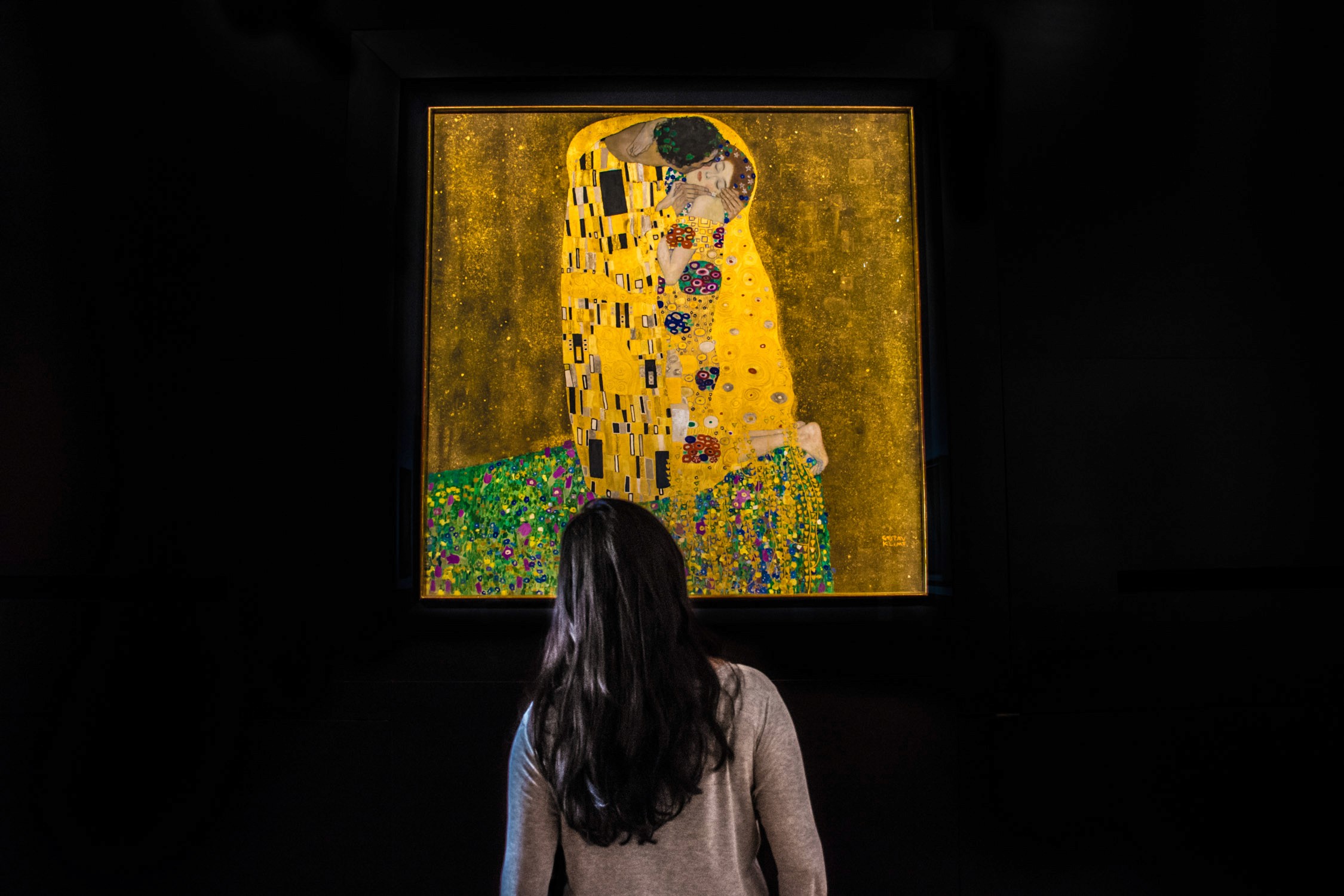 A person with long dark hair stands in a dimly lit room, viewing a large painting of a woman in a yellow dress.