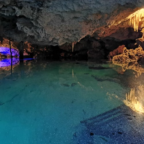 A brightly lit cave with stalactites reflecting in a clear, turquoise underground pool.