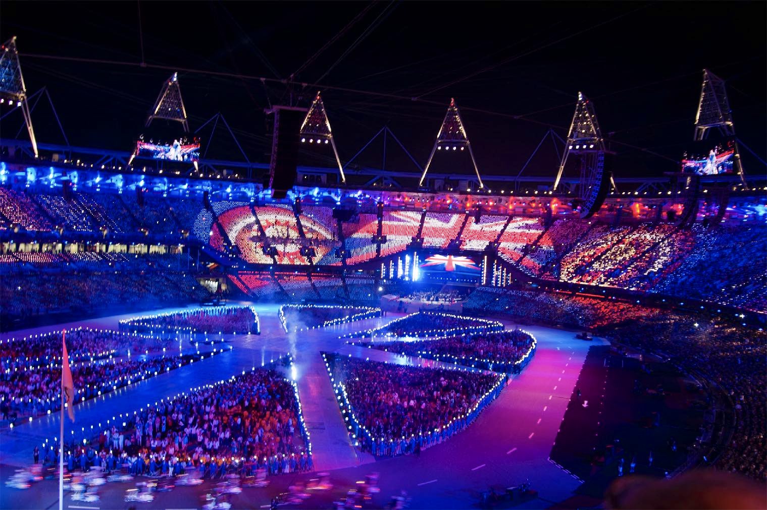 An illuminated stadium at night features vibrant, colorful lights and designs across the audience, creating a festive atmosphere during an event.