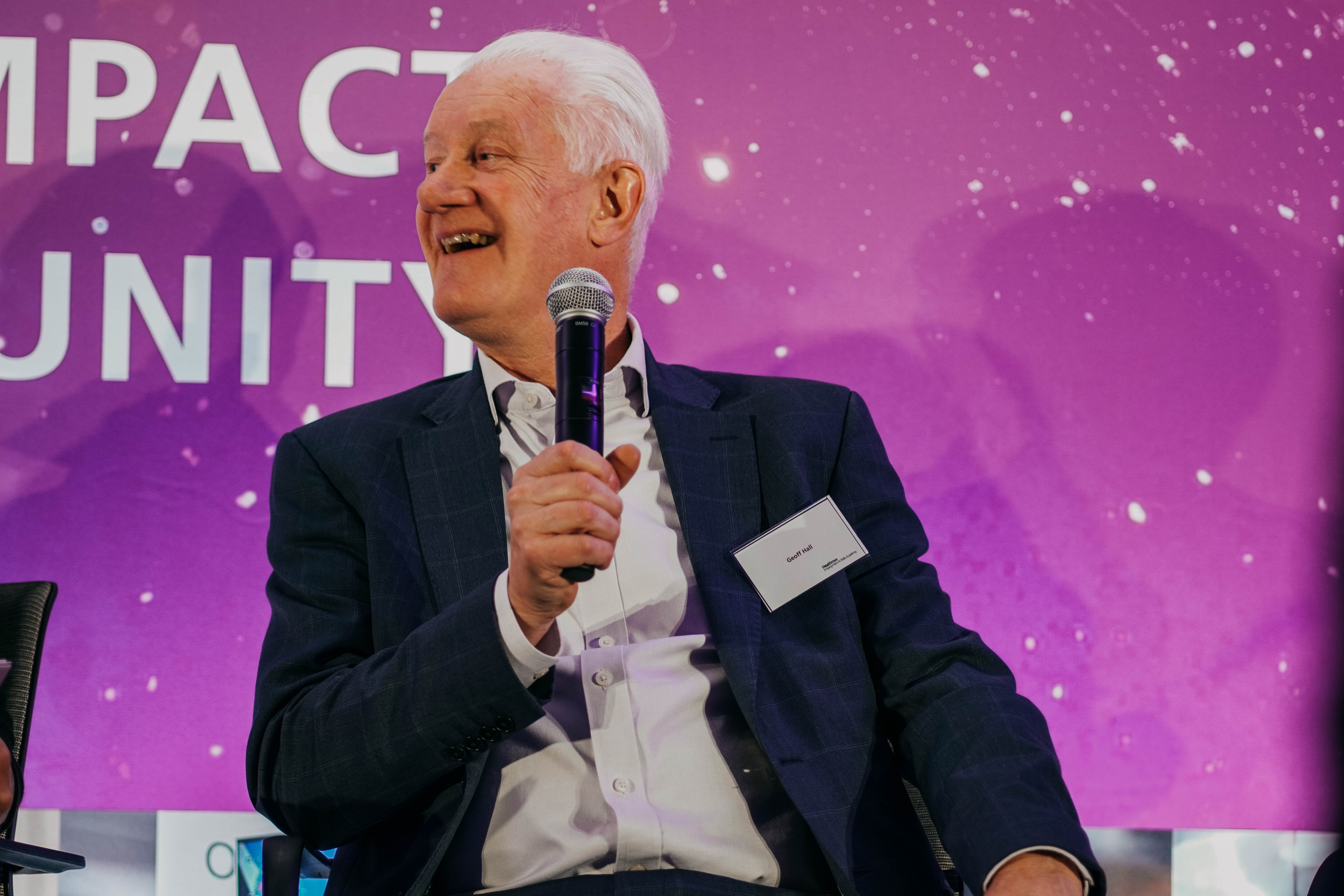 An older man with white hair, wearing a suit, smiles as he speaks into a microphone while seated on stage in front of a large purple backdrop.