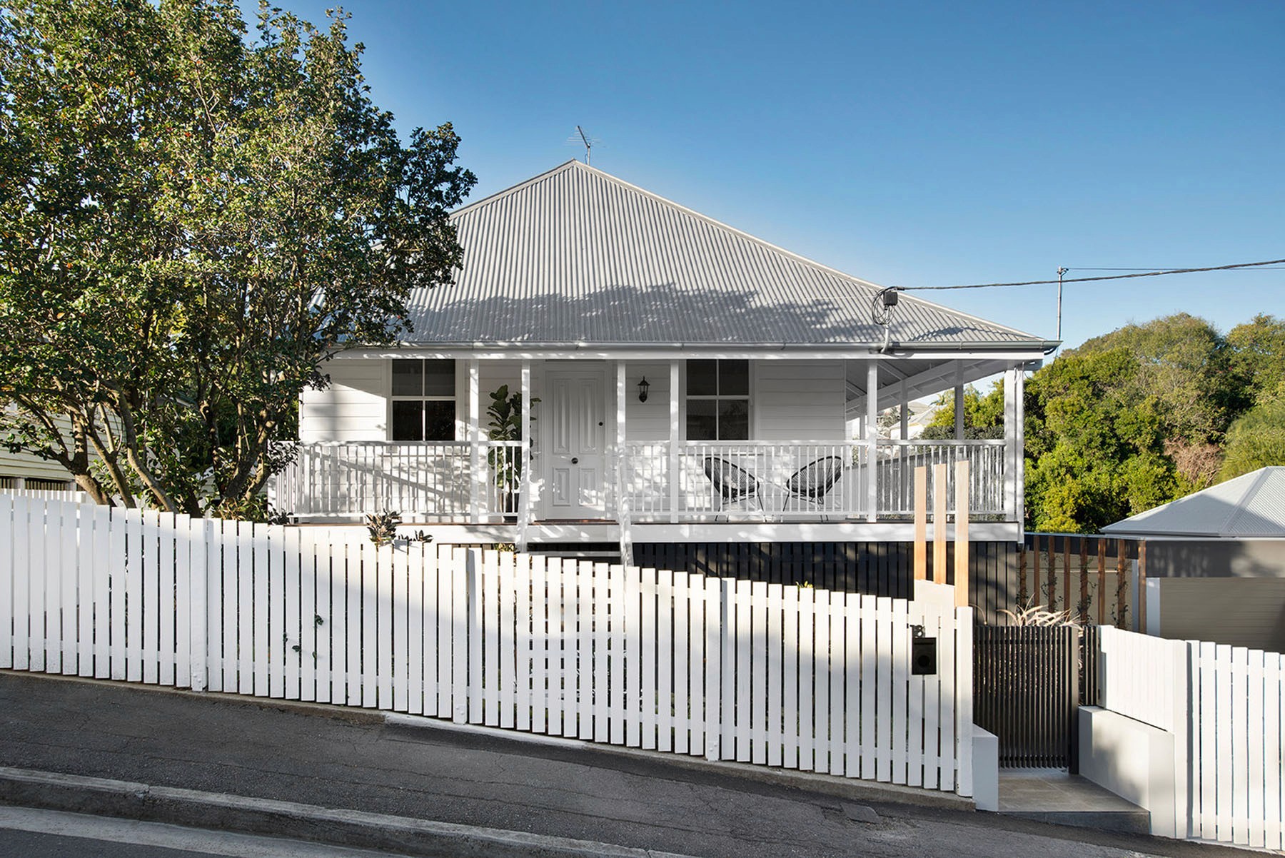 Restored Queenslander façade with corrugated roof, elevated veranda, and white picket boundary set against a sloping street.