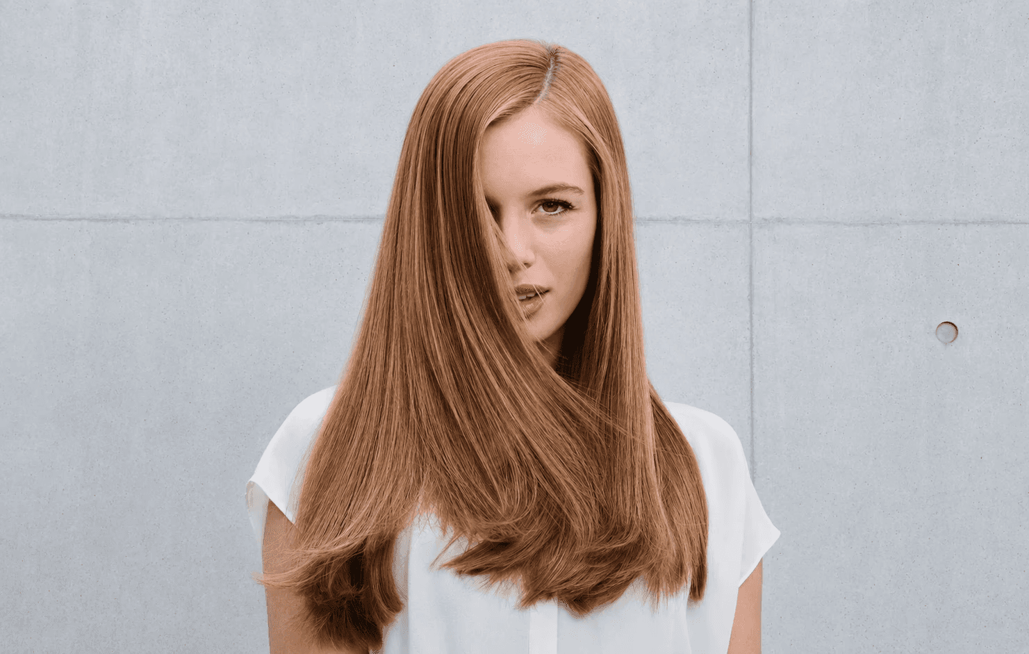 A woman with long, straight red hair stands in front of a plain background, facing the camera with a subtle expression.