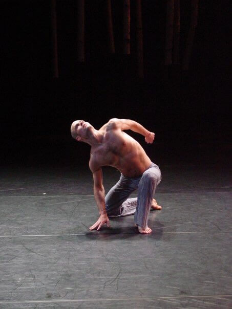 Mathew Janczewski, a shirtless contemporary dancer, kneeling on a dark stage, arching his back and looking upwards during a performance.