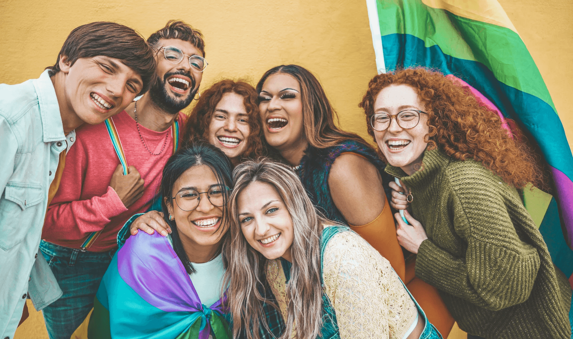 A group of seven diverse people smiling, posing together with a rainbow flag against a colorful background.