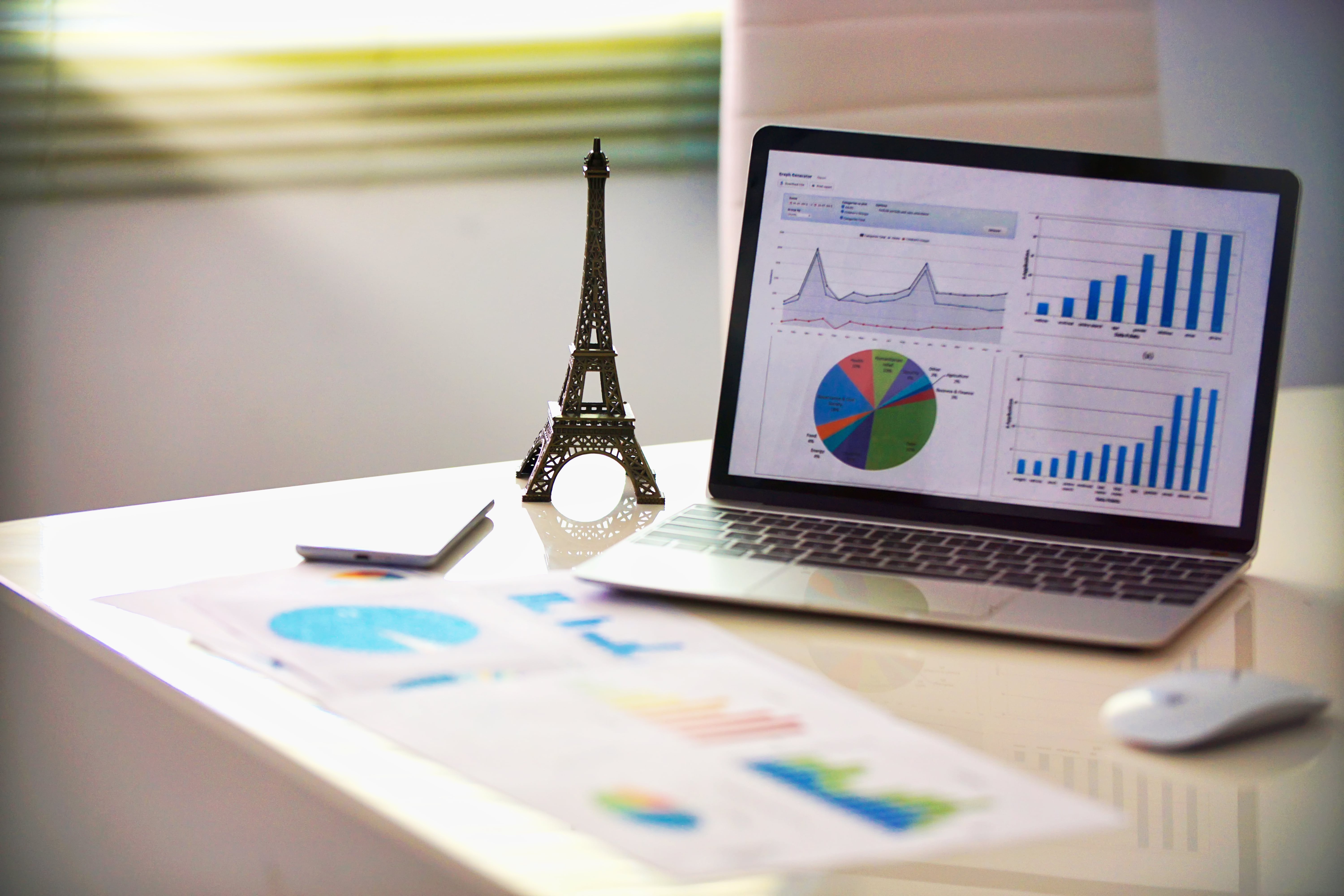 An office desk setup for business analytics, featuring a laptop displaying financial reports with charts and graphs. A miniature Eiffel Tower statue, a smartphone, and printed documents with data visualizations are on the desk, representing financial planning and global business.