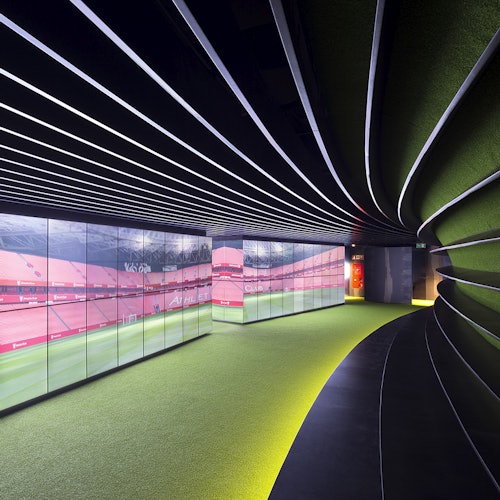 A modern hallway with curved walls, digital screens displaying red stadium images, and green flooring with overhead striped lighting.