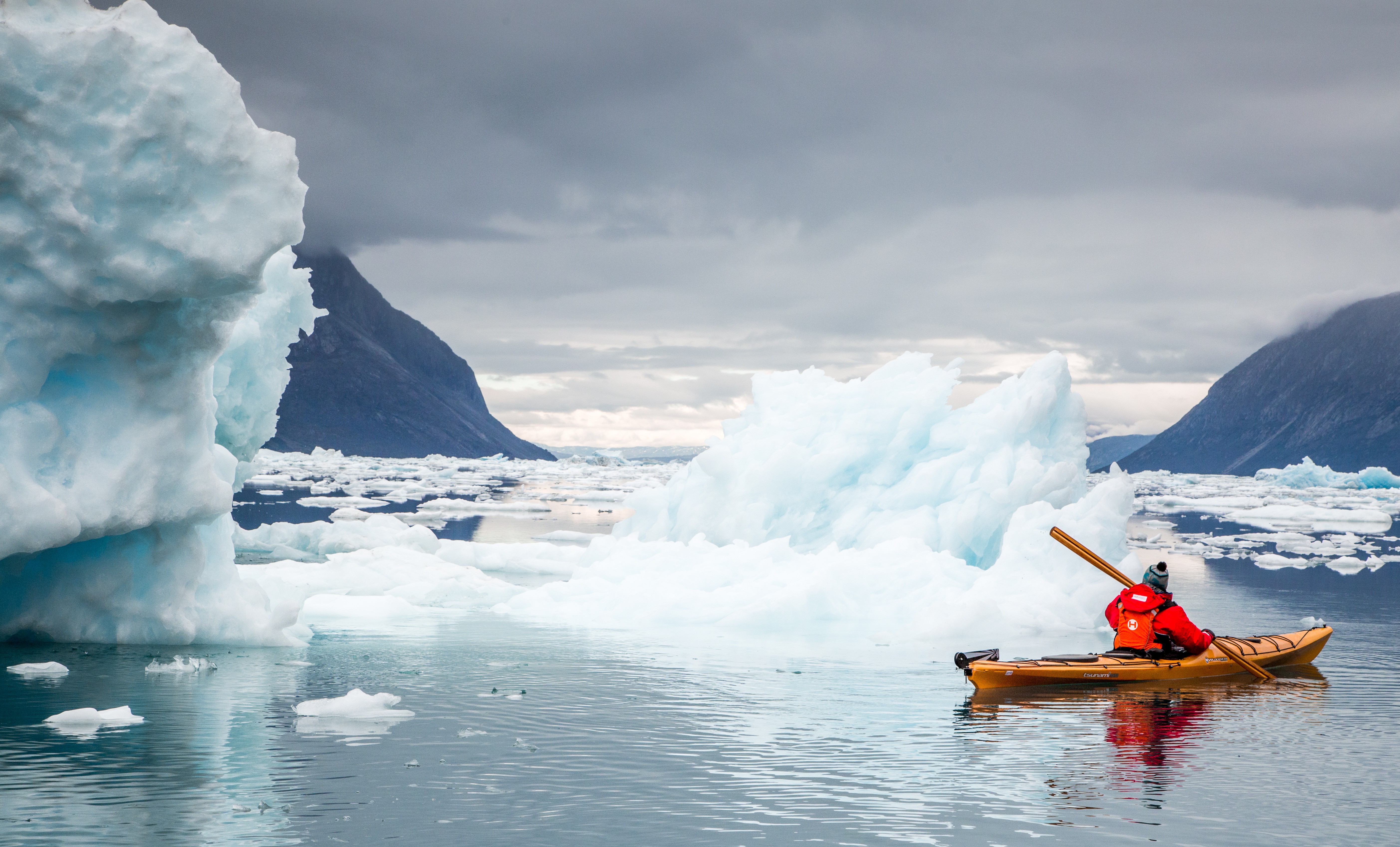 A kayaker in a red jacket paddles through icy waters surrounded by floating icebergs and towering mountain cliffs.
