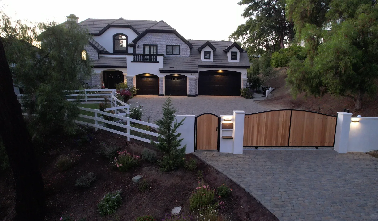 Paver driveway and wooden gate framed by planting beds and a white fence in front of a large house at dusk.