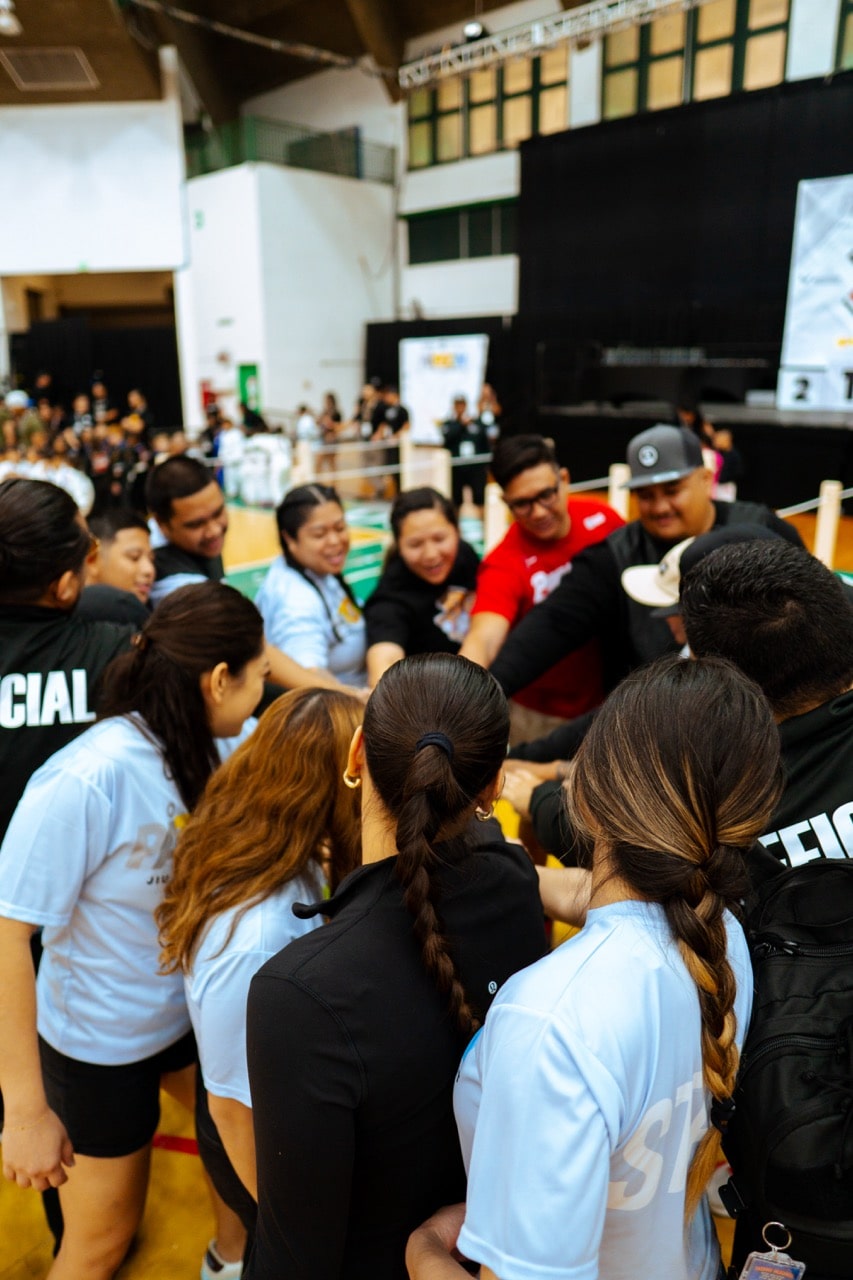 A group of event organizers and staff members stand in a huddle, with their hands in the center, showing teamwork and solidarity at a jiu-jitsu tournament