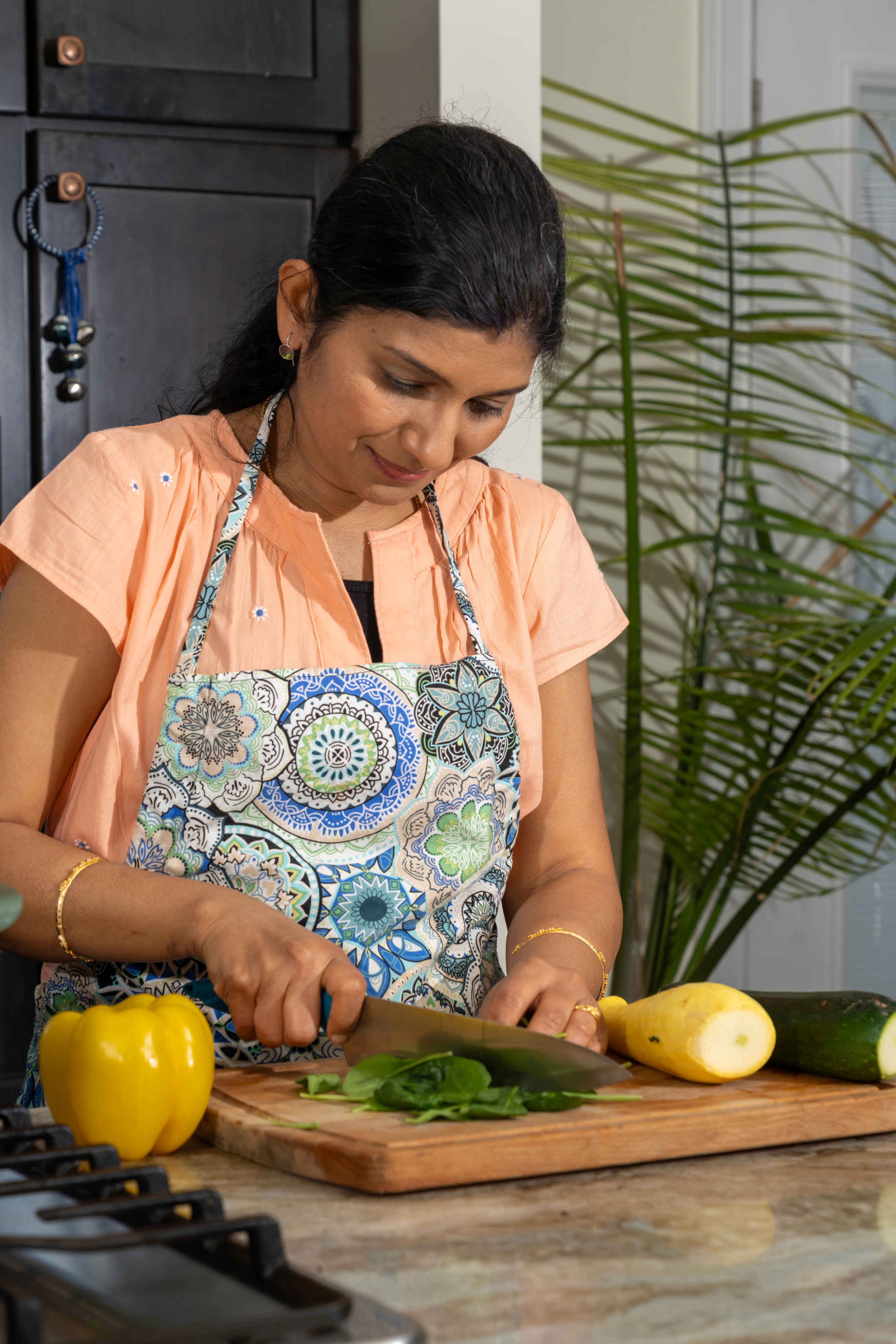 A woman in a floral apron chops fresh greens on a wooden cutting board in a bright kitchen, surrounded by a yellow bell pepper, zucchini, and vibrant plant, reflecting a healthy lifestyle. Ayurvedic diet consultation Louisville
