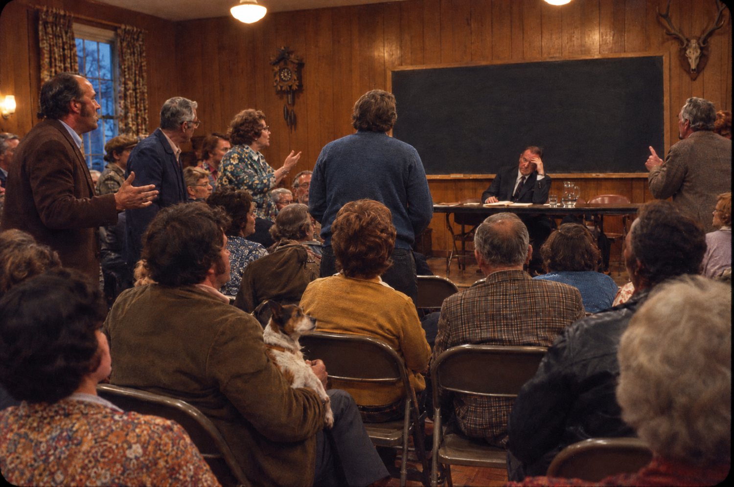 A crowded, arguing town meeting in a wood-panelled hall, a man with a small dog on his lap, the mayor with his head in his hand