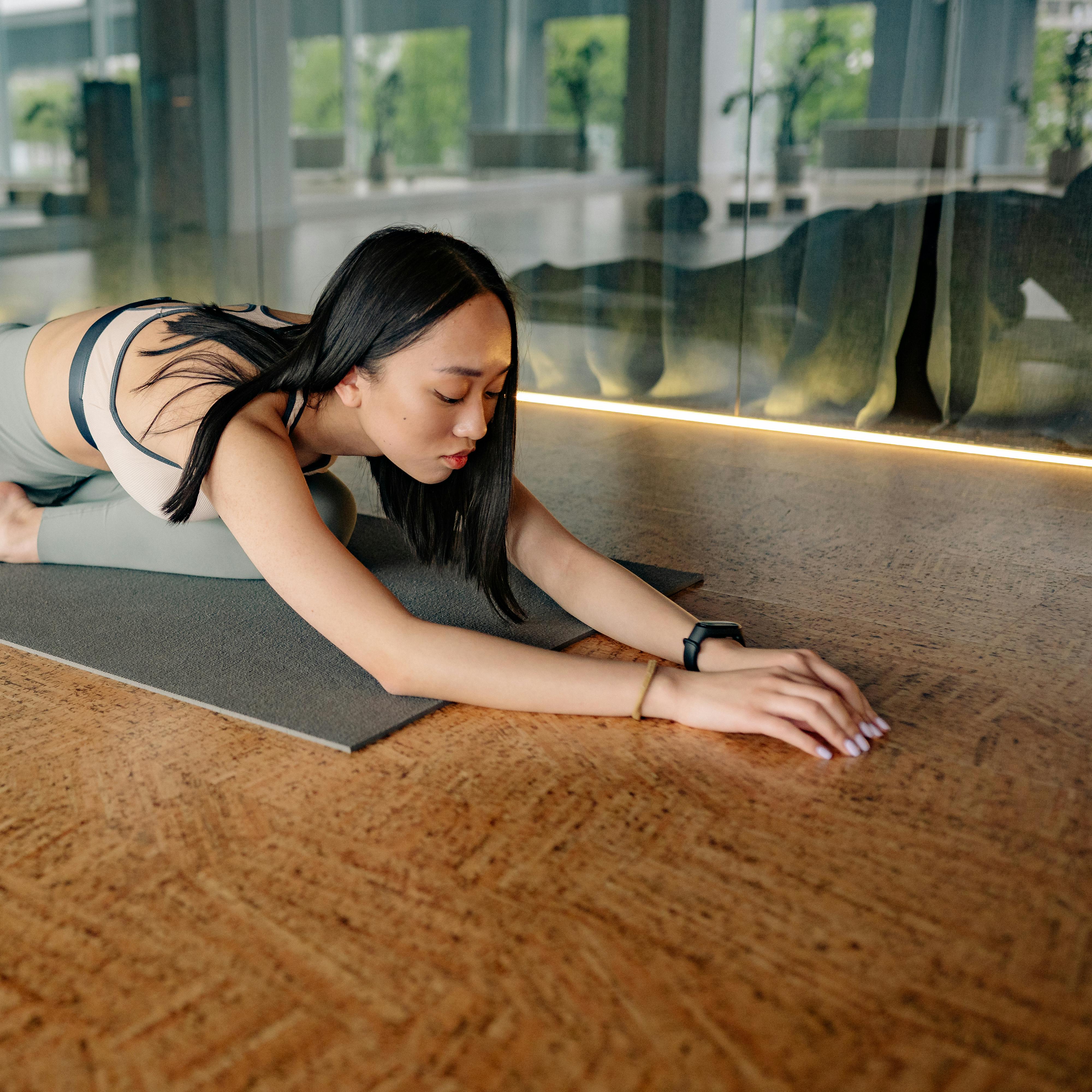 A woman practices yoga indoors, stretching on a mat in a peaceful position. The room features large windows with greenery visible outside, conveying calmness.