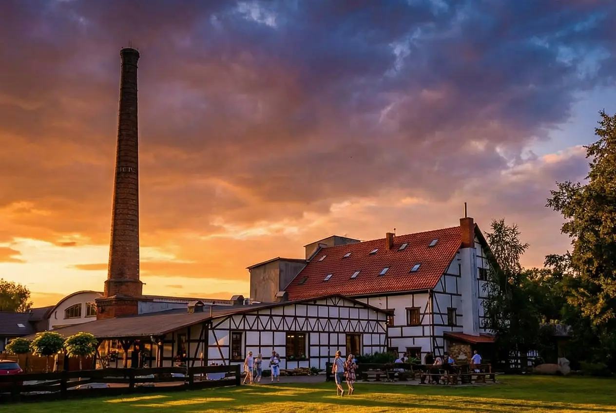 Zachód słońca nad Wrzosowym Tartakiem, z widocznym kominem i oświetlonym budynkiem w otoczeniu zieleni.