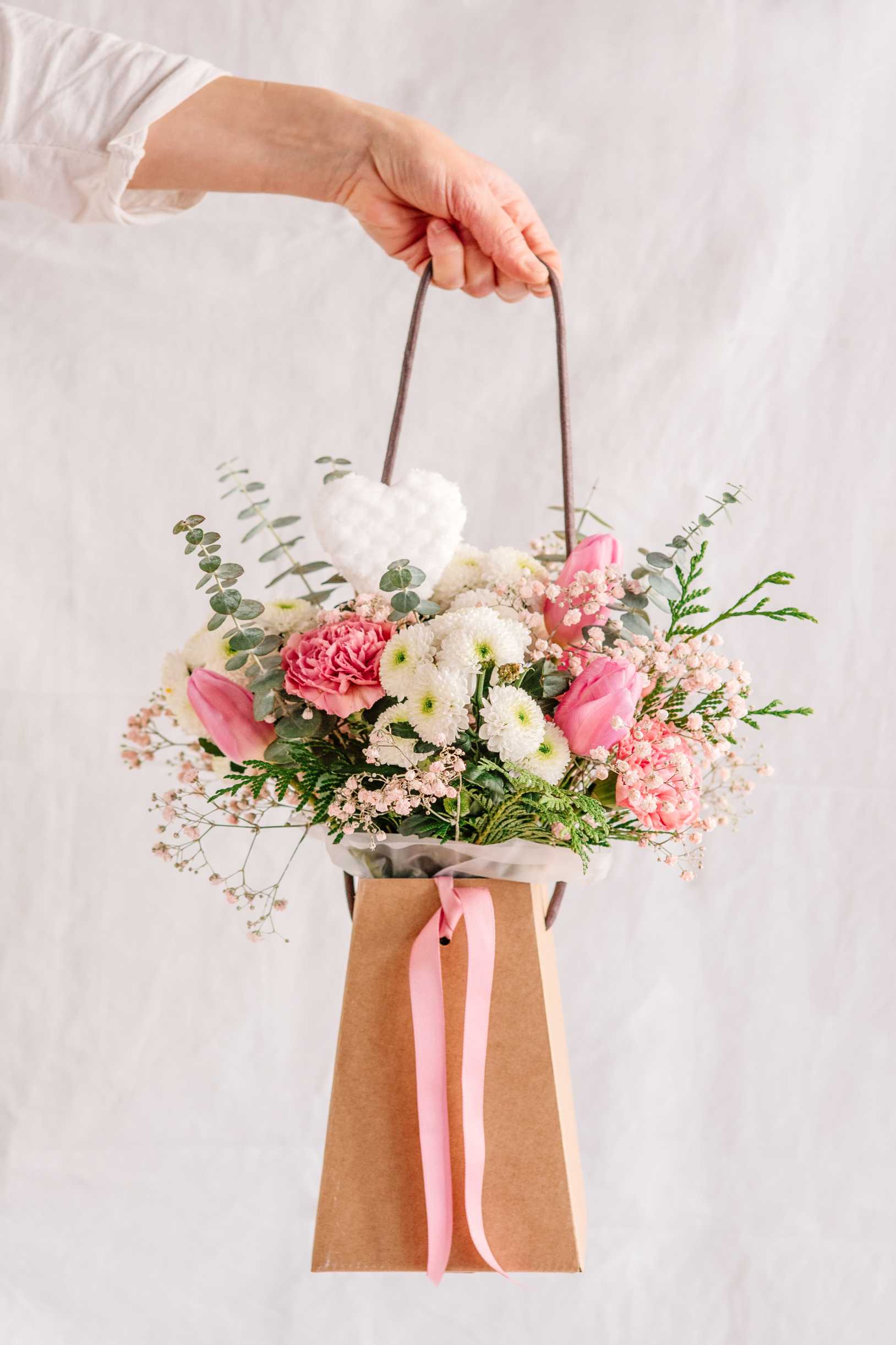 Bouquet floral en tons romàntics subjectat per una mà, presentat dins una bossa kraft amb nansa i cinta rosa.