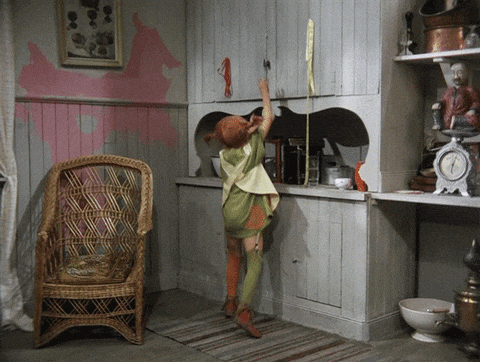 A child wearing a colorful outfit with green and orange hues reaches up to open a cabinet in a rustic kitchen, which features painted cupboards, a wicker chair, and vintage decor, adding to the quaint atmosphere.