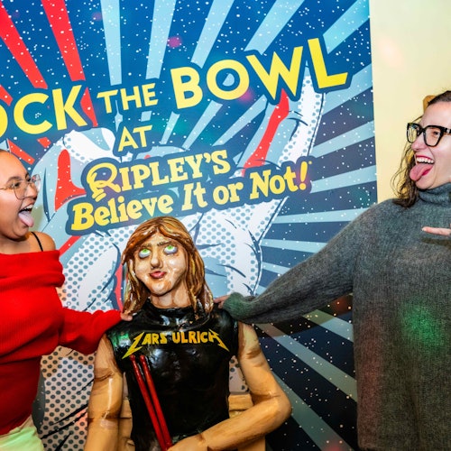 To personer griner og poserer med en Lars Ulrich-figur på Ripley's Believe It or Not, foran en farverig stjerneskudsbaggrund.