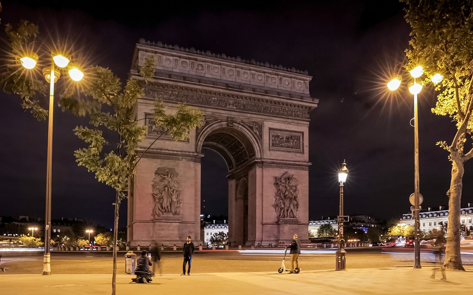 巴黎的凯旋门在夜晚灯火辉煌,游客与滑板车穿梭其中。