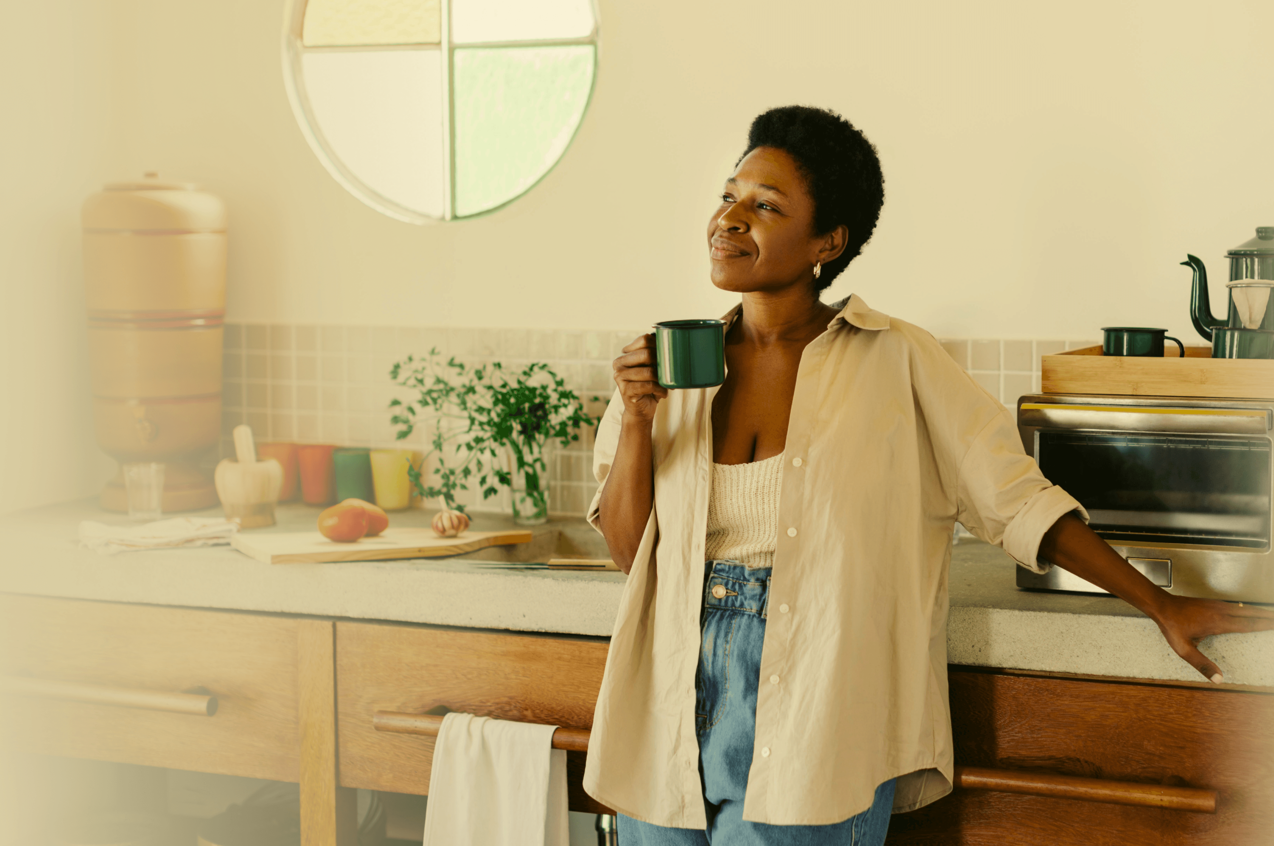 Mulher negra olhando para a janela do seu apartamento tomando um café na sua caneca de cor verde.