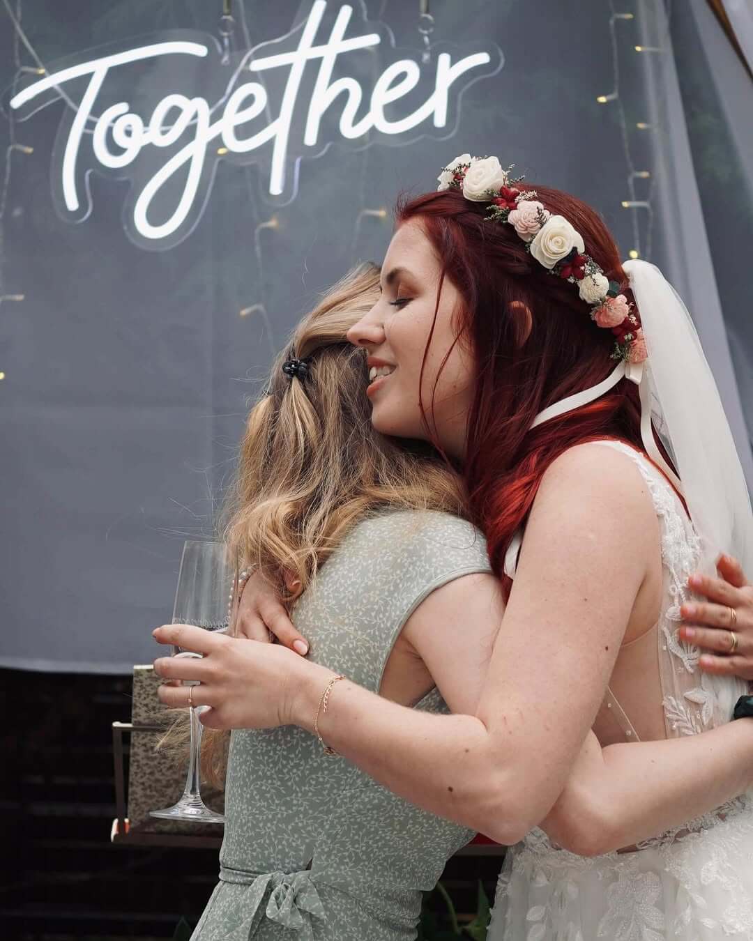Zwei Frauen umarmen sich vor einer Neon-Schrift "Together". Eine trägt ein Brautkleid mit Blumenkranz.