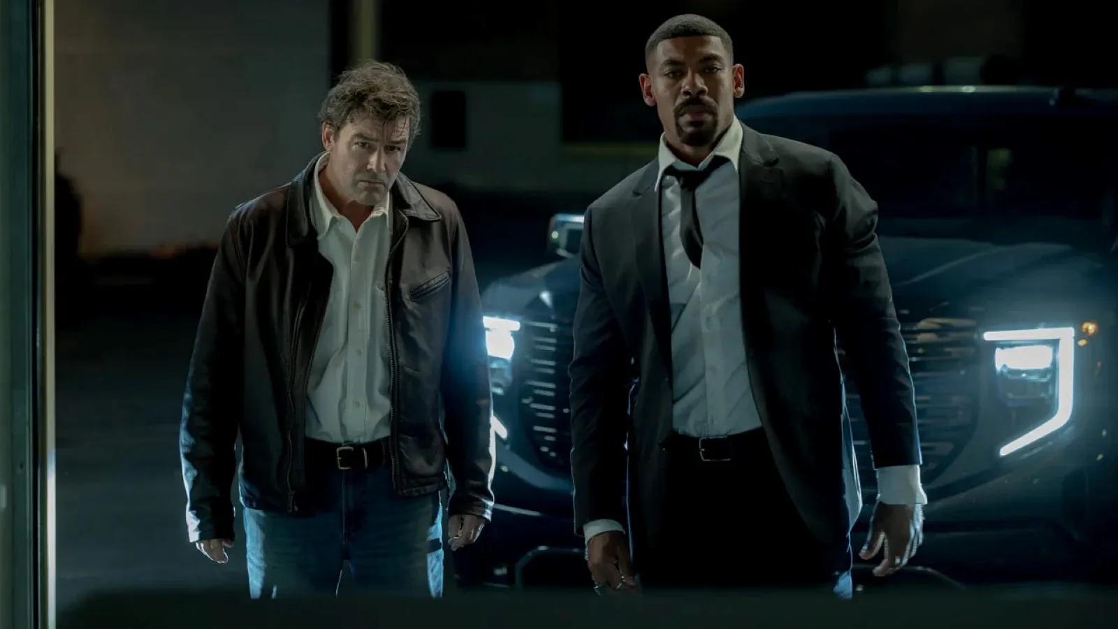 Two men stand in front of a dark SUV with illuminated headlights, one wearing a leather jacket and the other in a suit and tie, looking intently towards the camera.