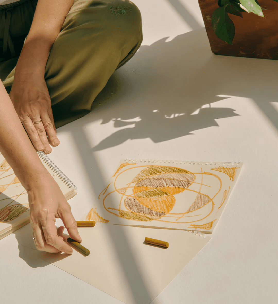 Girl sitting on the floor, drawing with orange and brown crayons on white paper, symbolizing the importance of mindfulness in mental and emotional wellbeing.