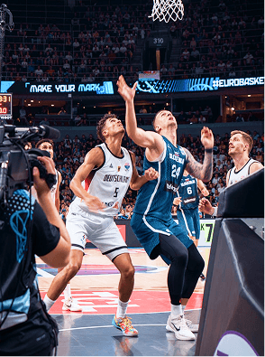 Basketball players jump for the ball during a game. One player wears a German jersey and another wears a blue jersey.