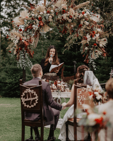 Ein Brautpaar sitzt bei einer freien Trauung im Freien unter einem opulenten Blumenbogen. Die Braut trägt ein weißes Kleid mit Schleier, der Bräutigam einen dunklen Anzug. Sie halten sich liebevoll an den Händen. Vor ihnen steht eine Traurednerin mit Mikrofon und Buch, während sie feierliche Worte spricht. Die Szenerie ist von Natur umgeben und wirkt romantisch und stilvoll dekoriert.Maria Wiethaler freie Rednerin und Sängerin für Hochzeiten, Taufen, Trauerfeiern und Events aus München und Landshuter Umgebung