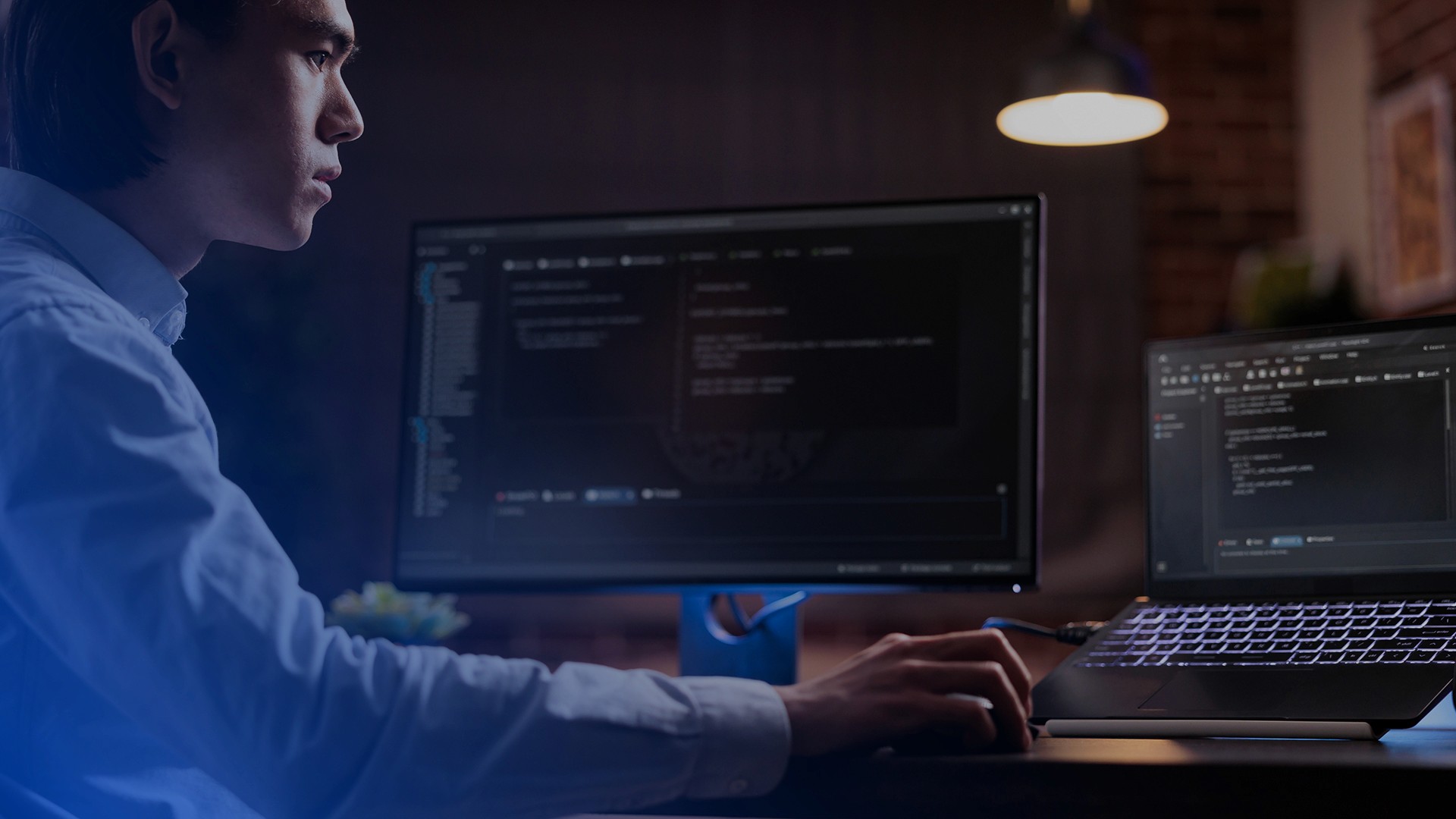 A person in a light-colored shirt sits at a desk in a dimly lit room, working on both a laptop and external monitor. The screens display lines of code or programming text, and the scene is illuminated with blue-tinted lighting. A pendant lamp is visible in the background.