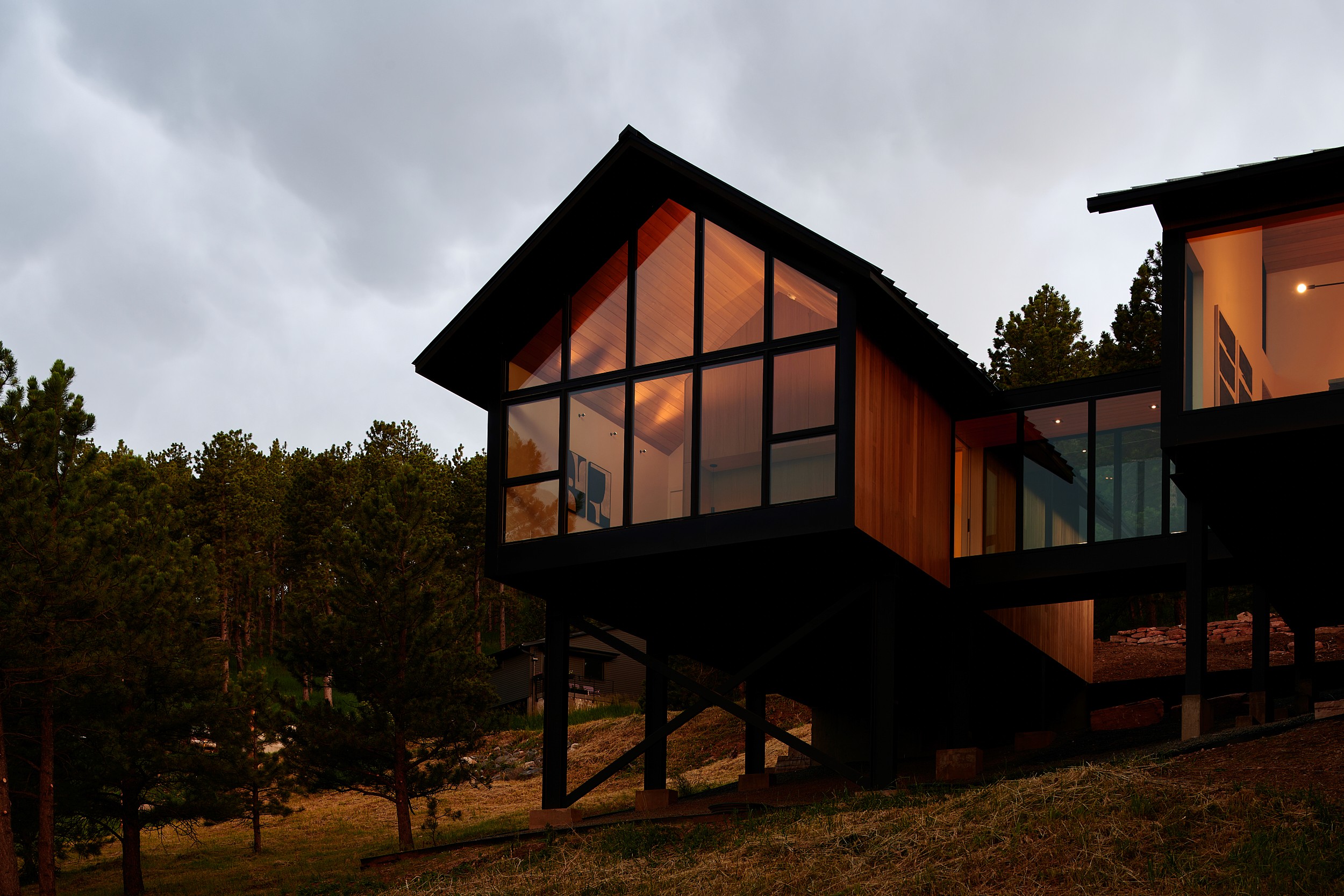 A modern mountain home with glass bridge and gable form with glass end wall floating over the landscape on steel structure with, glowing from the interior at evening.