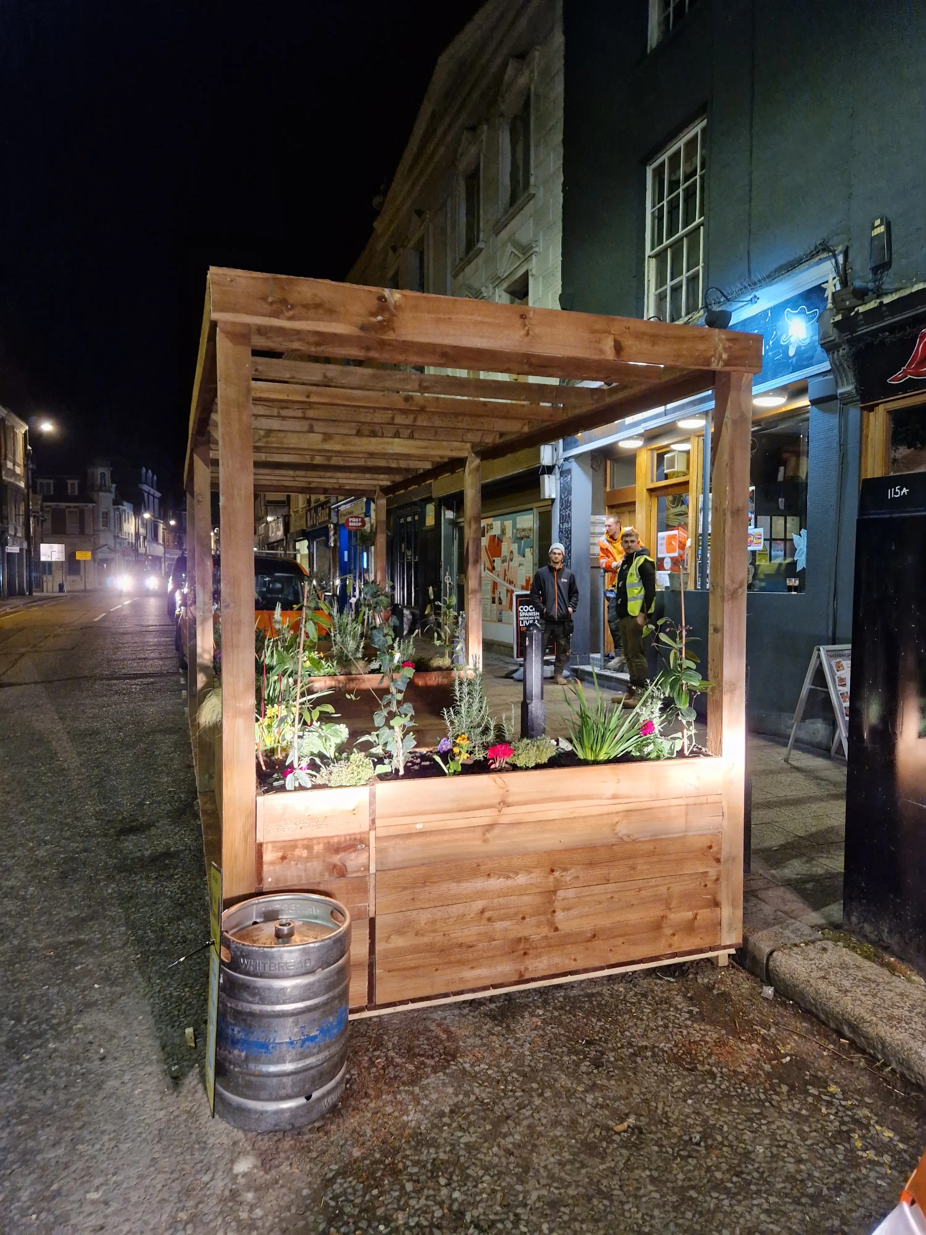 A wooden stall filled with plants and flowers, illuminated at night on a street with nearby buildings.