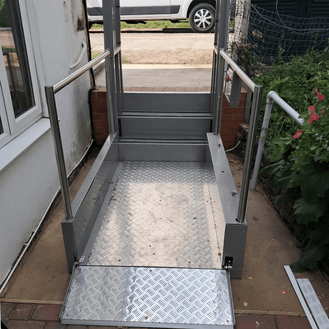 An outdoor vertical platform lift installed at a UK residential property, showing the open checker-plate platform, stainless steel safety rails, and automatic entry ramp at ground level.