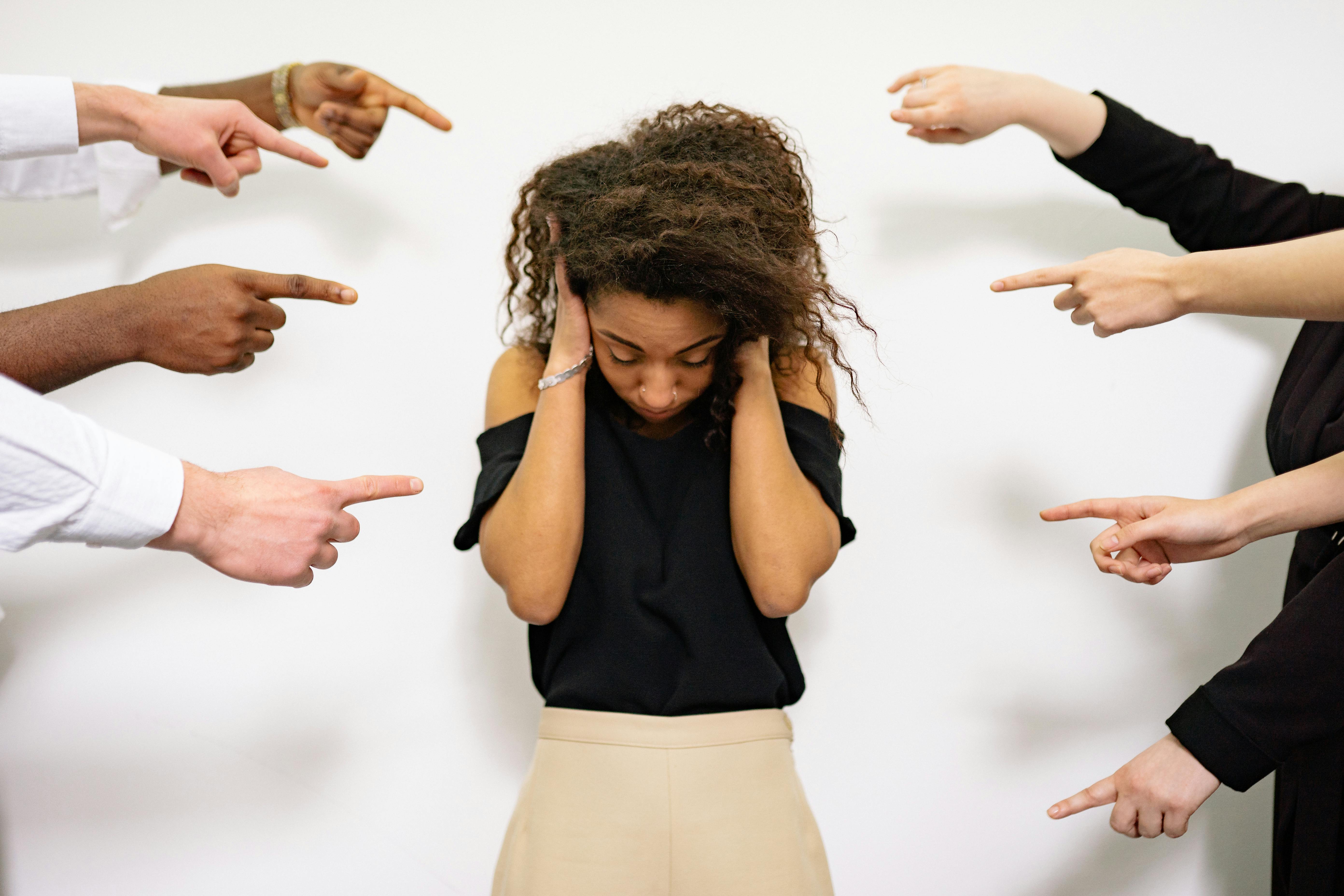 A woman is standing nervous in the middle of a group with 8 fingers pointed at her. She is visibly nervous, covering her ears and looking down while closing her eyes. Her whole body is tense and shoulders lifted