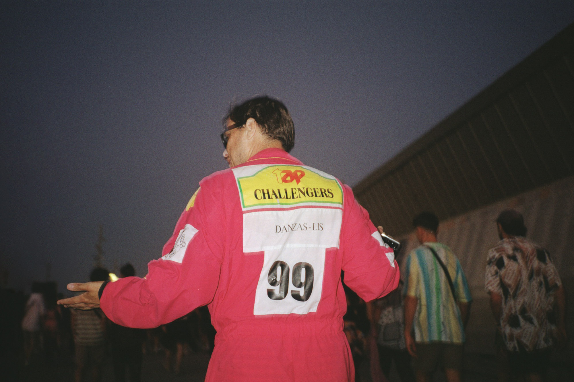 A person wearing a bright red racing jumpsuit with the words "Challengers" and the number "99" printed on the back stands outdoors in a crowd, with a dusky sky and a modern building in the background.