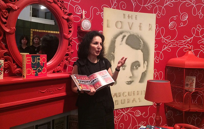 A photo of a woman giving a lecture and holding an opened book while sitting in a room with red walls and other red furniture.