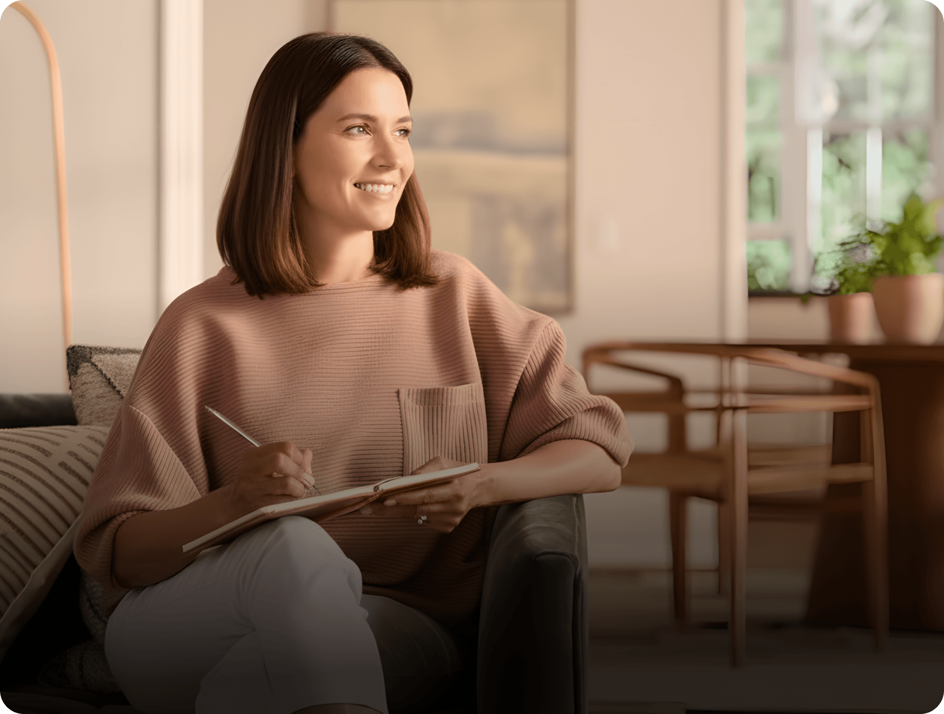 Trade name availability search - Woman sits on a couch in a living room, smiling and looking out a window while holding a pen and notebook.